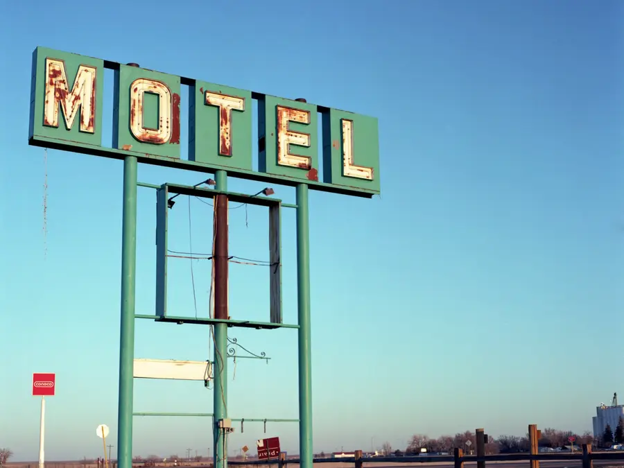 An abandoned roadside motel sign on the plains at dusk: rusted neon letters on a faded green panel, the frame below it stripped bare, a Conoco sign in the distance.